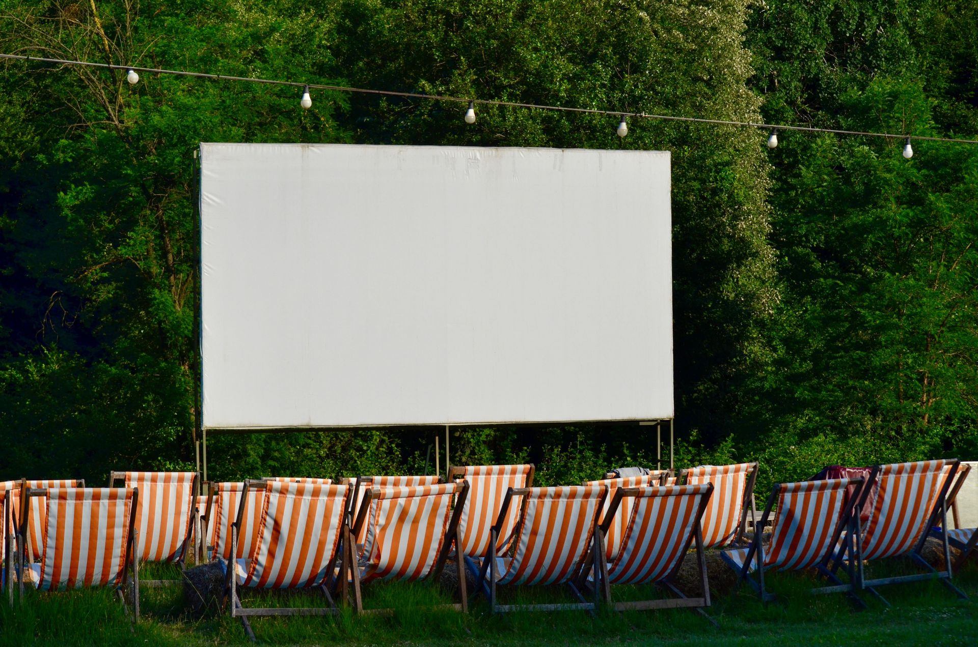 Empty outdoor cinema venue with striped deck chairs and a blank screen, surrounded by greenery.